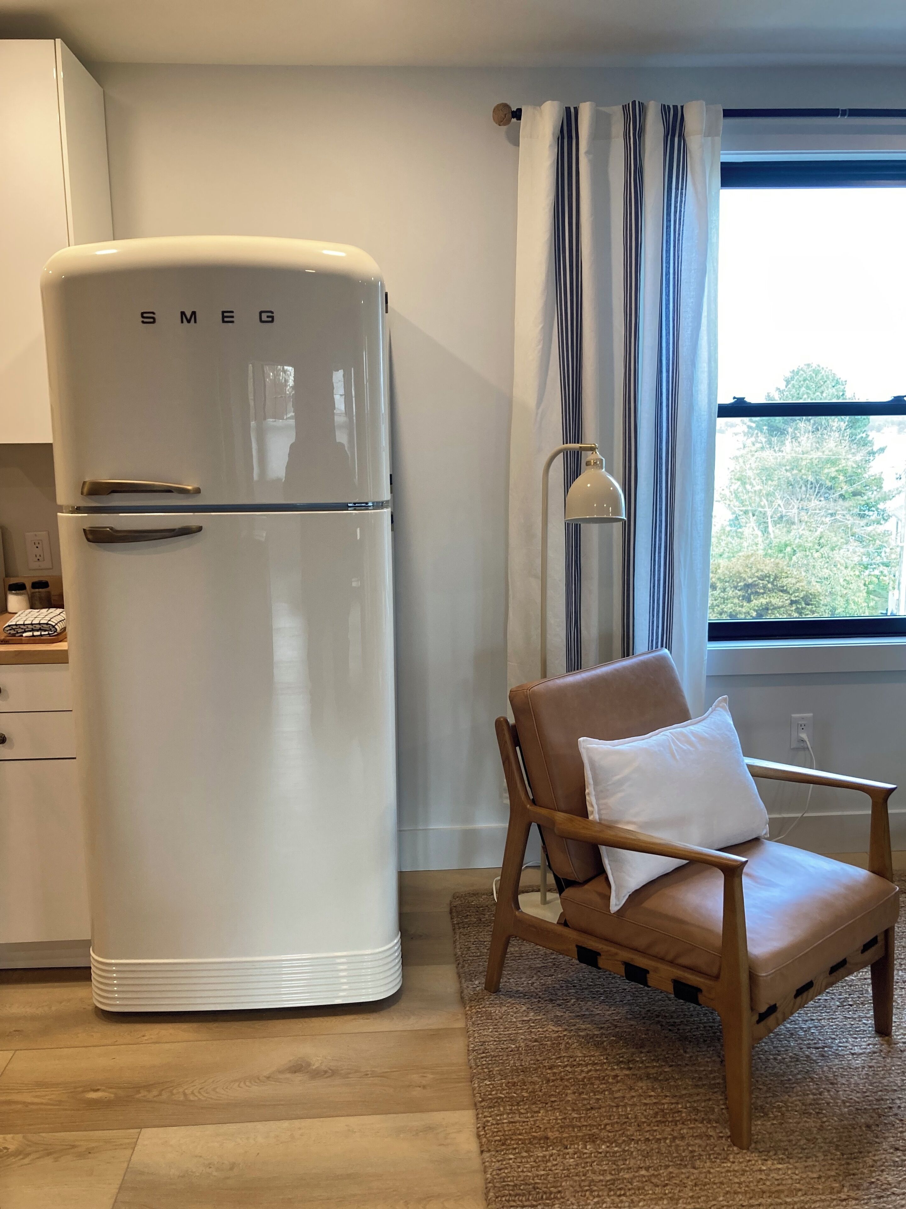 A compact kitchen with a white Smeg refrigerator, a wooden chair with a white cushion, and a window with curtains.