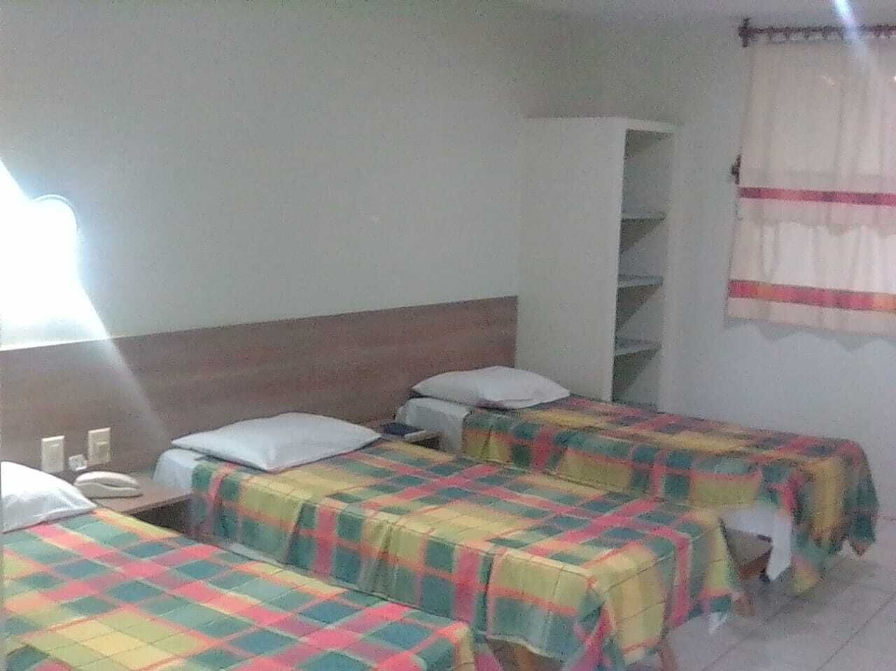 Una habitación con dos camas, cada una con una colcha colorida, una pared blanca, una ventana con cortinas y una mesita de noche con una lámpara.