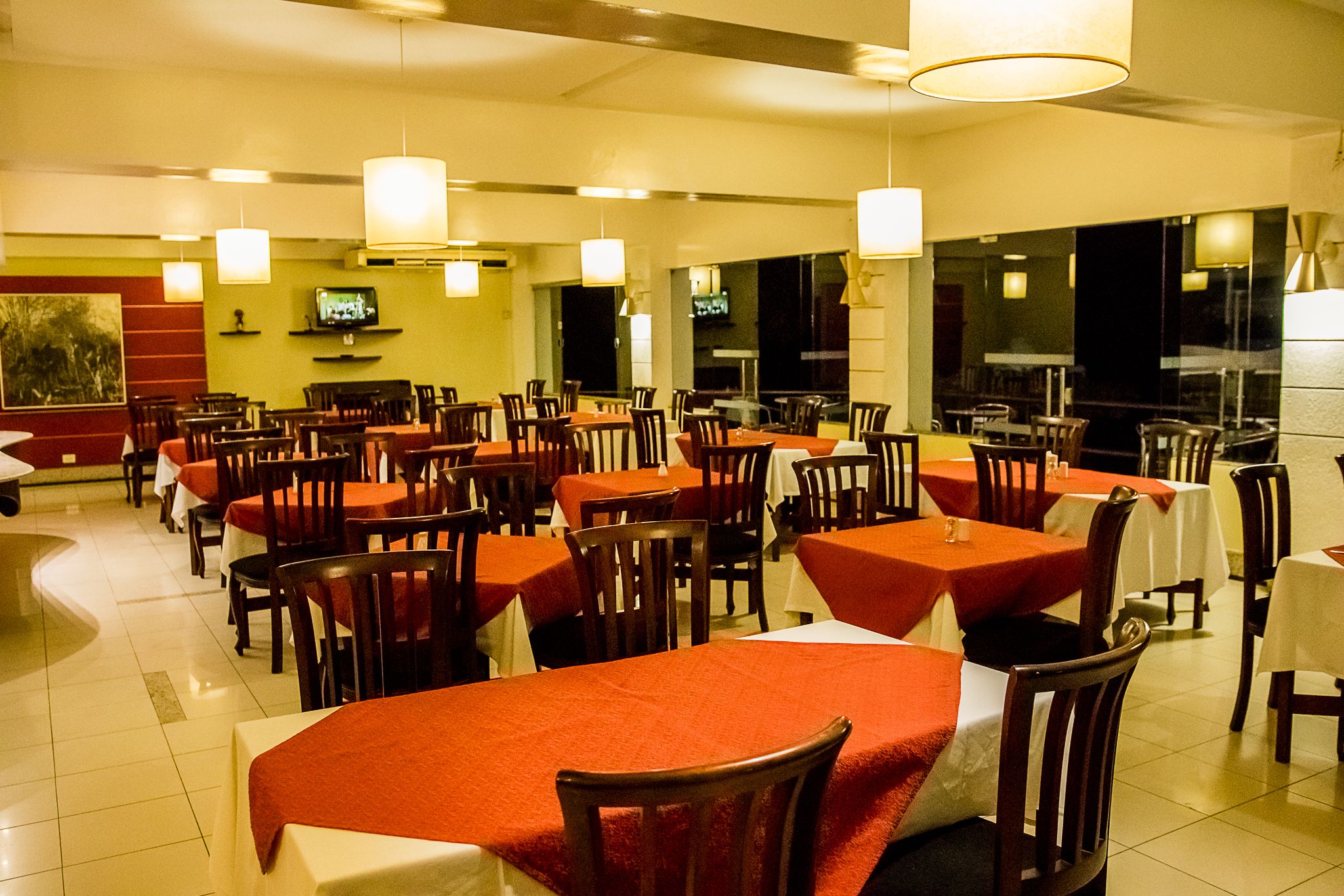 A restaurant with multiple tables and chairs, overhead lighting, and a television on the wall.