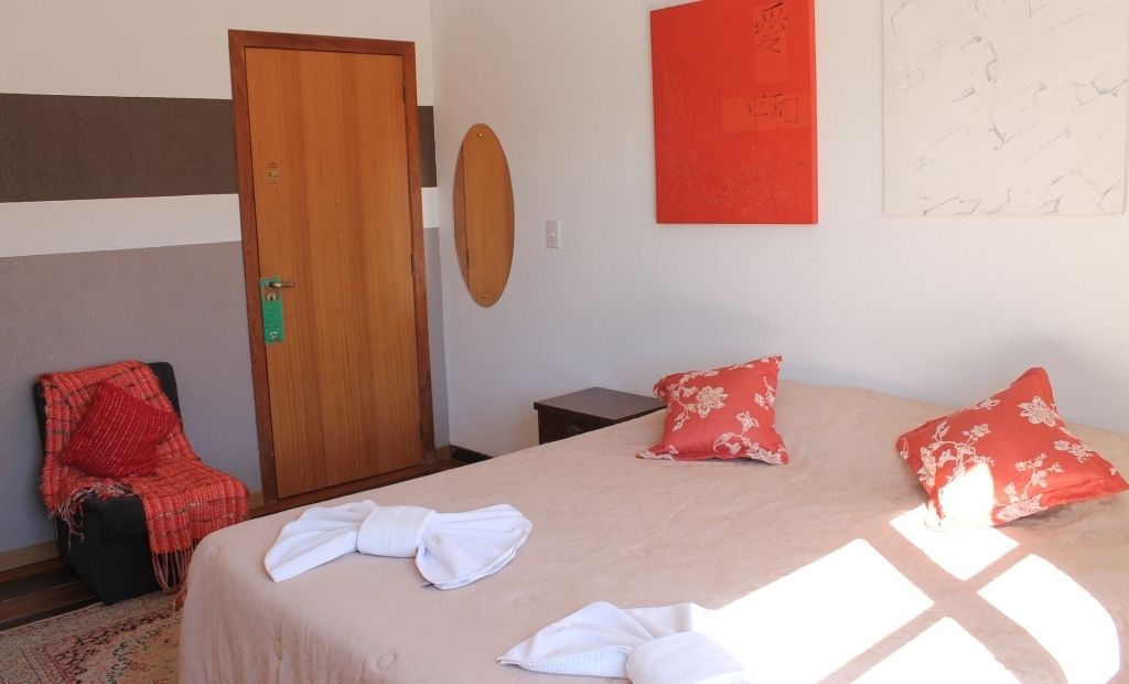 Quarto de hotel com cama, duas almofadas, uma cadeira, porta de madeira e decoração vermelha na parede.