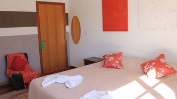 Quarto de hotel com cama, duas almofadas, uma cadeira, porta de madeira e decoração vermelha na parede.