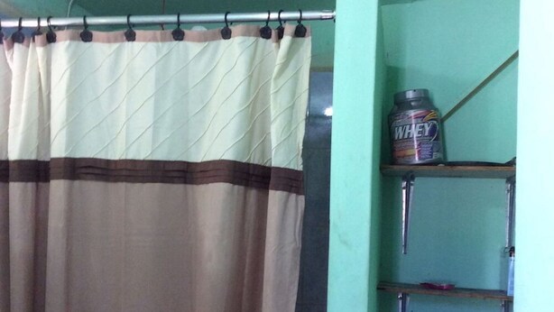 A bathroom with a beige and brown shower curtain, a WHEY protein powder shelf, and a metal pipe. - MaFlo Arms Apartments (Belize City)
