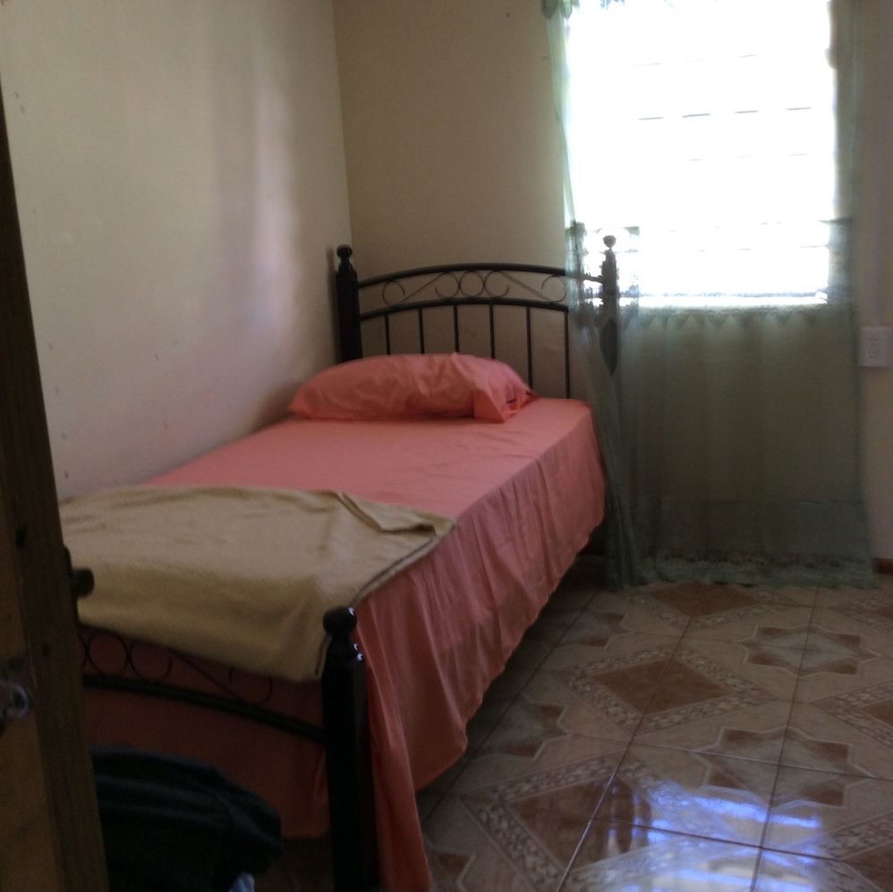 Een kleine kamer met een enkel bed, een metalen bedframe, een roze sprei en een beige deken. De vloer is betegeld met een geometrisch patroon.