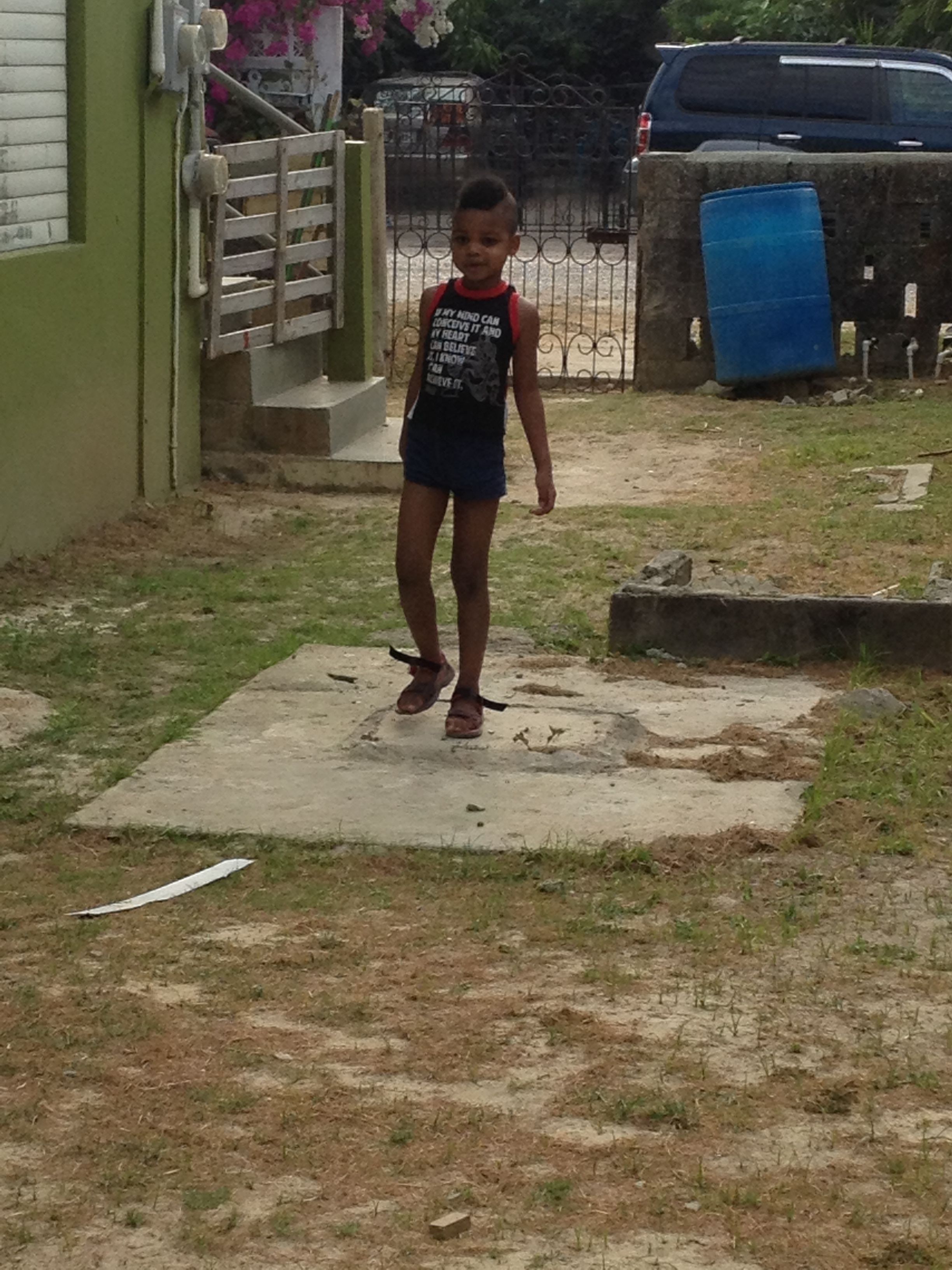 Un niño de pie sobre una losa de hormigón en un patio con un muro verde y una valla metálica al fondo.