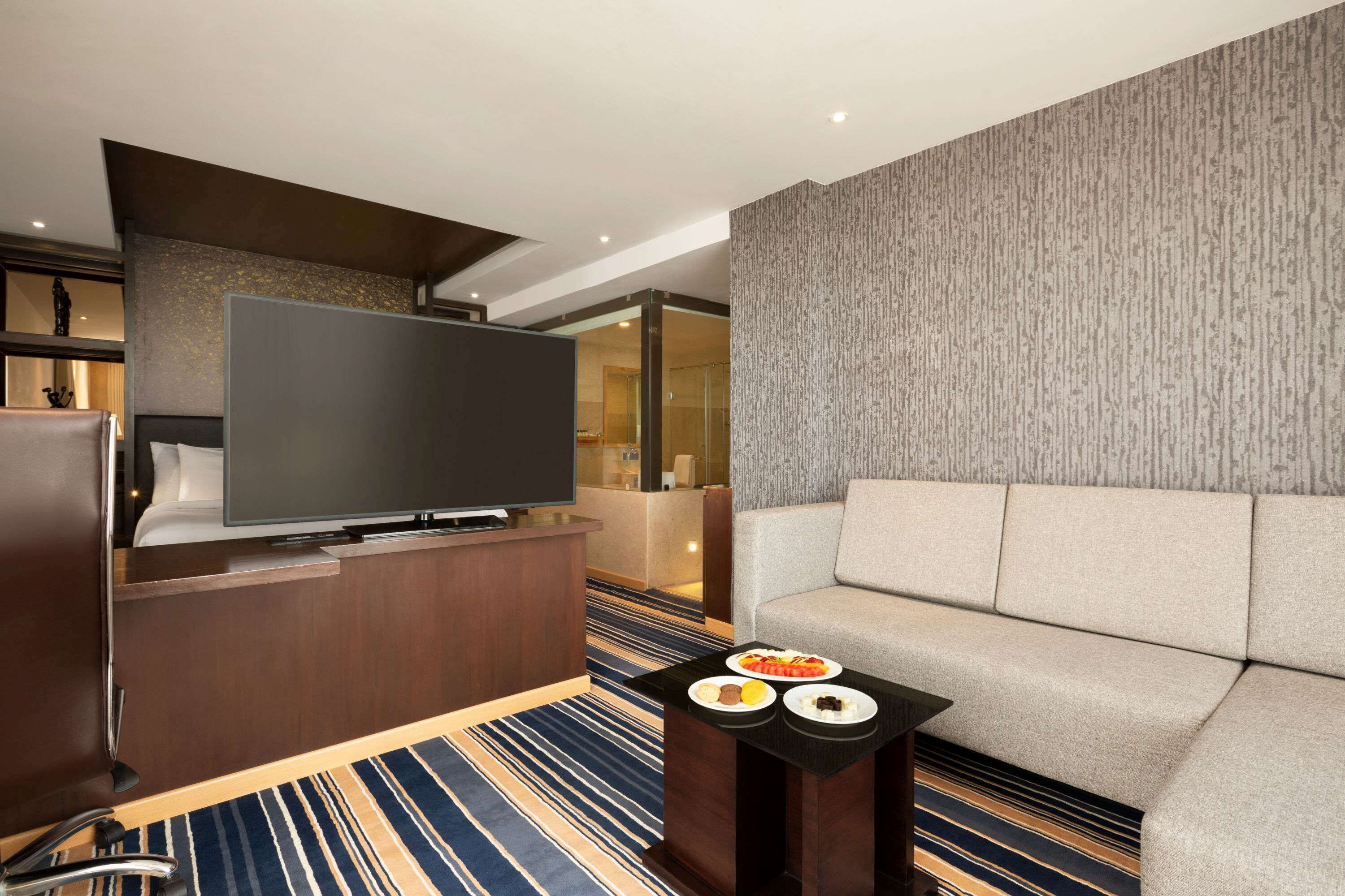 A modern hotel room with a large flat-screen TV, a sofa, a small table with plates of food, and a striped carpet.