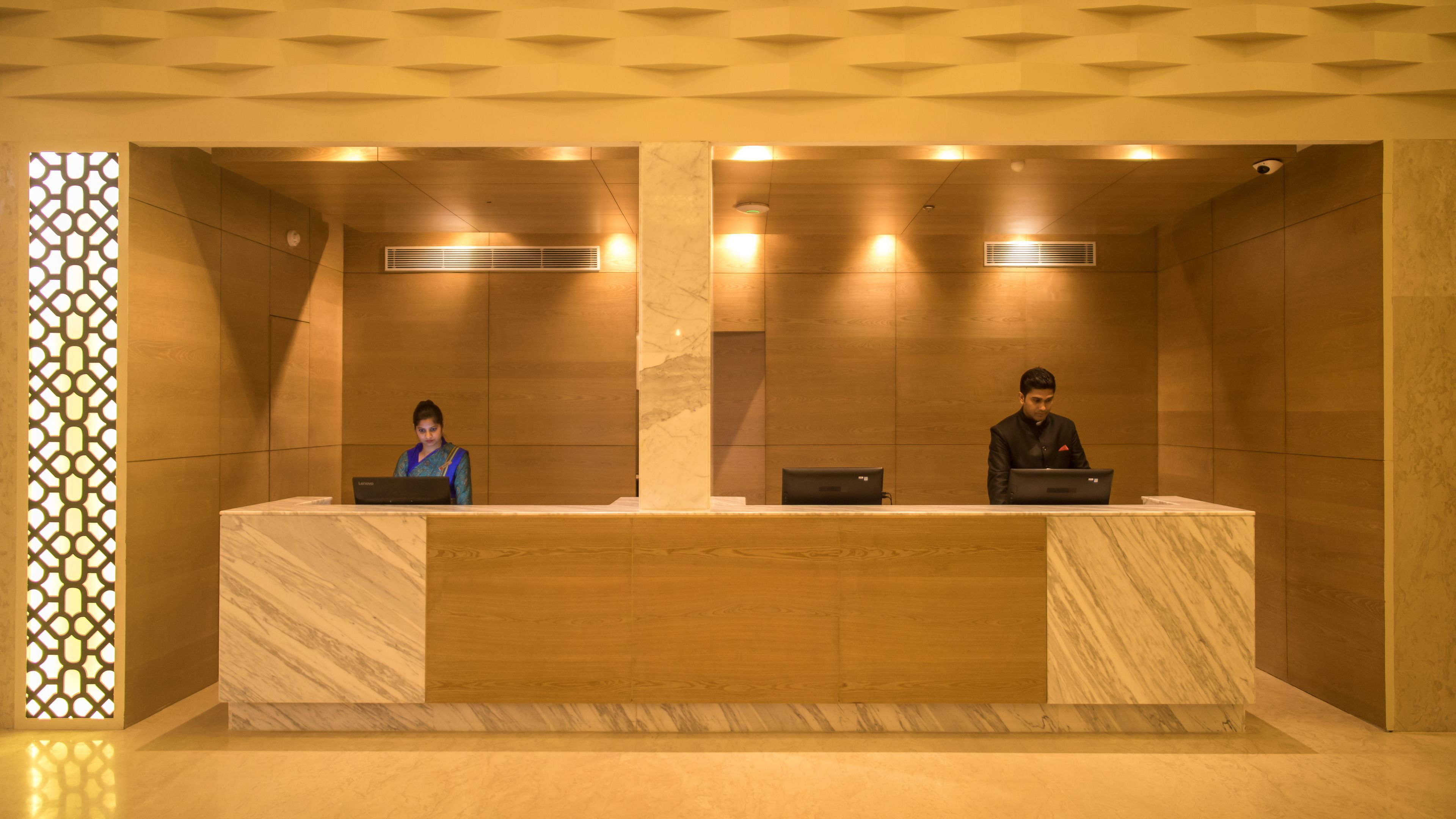 A reception desk with two staff members, a patterned wall, and a ceiling with recessed lighting.