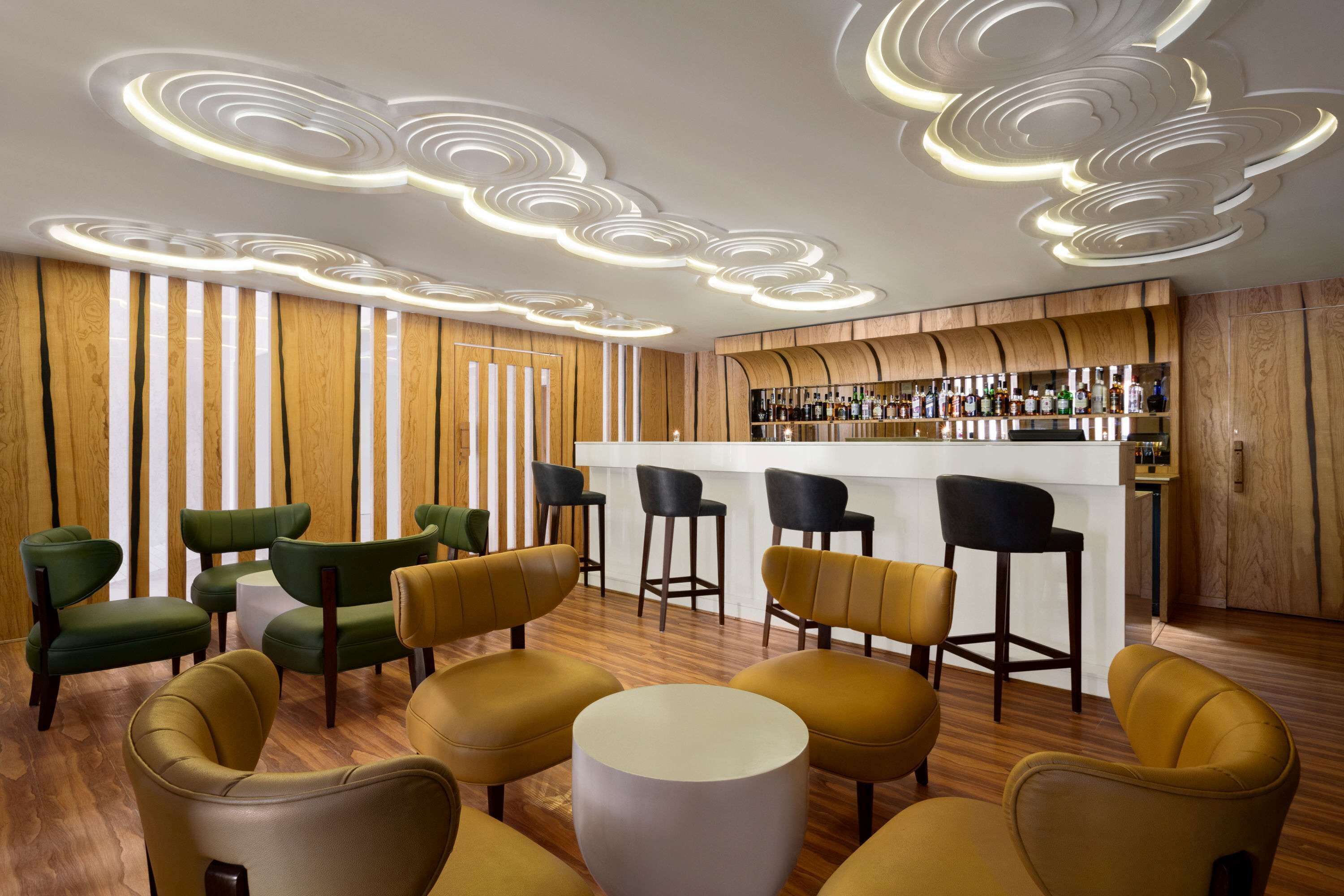 Een bar met een houten toog en krukken, omringd door groene en gele stoelen.