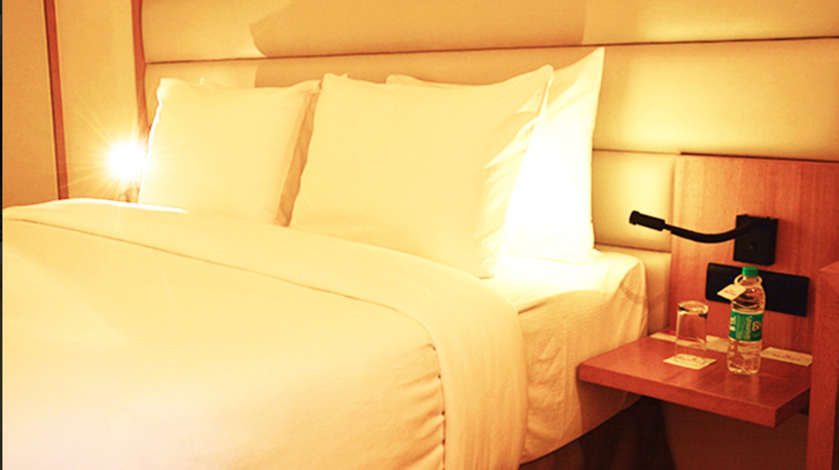 A hotel room with a bed, pillows, a nightstand with a bottle of water, and a lamp.