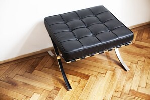 Un pouf en cuir noir, doté d’un motif capitonné, de pieds en métal et d’une base en bois, posé sur un parquet à motifs en chevrons.
