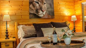 Ein Schlafzimmer in einer Holzhütte mit einem großen Katzenbild, einem hölzernen Bett mit Kissen, einem Nachttisch mit Lampe und einem Tablett mit einer Weinflasche und einem Glas.