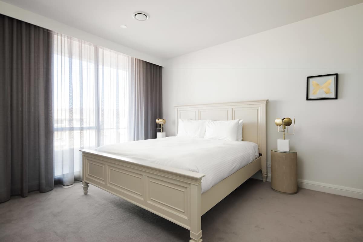 Un dormitorio con una cama grande, una mesita de noche, una lámpara y una ventana con cortinas.