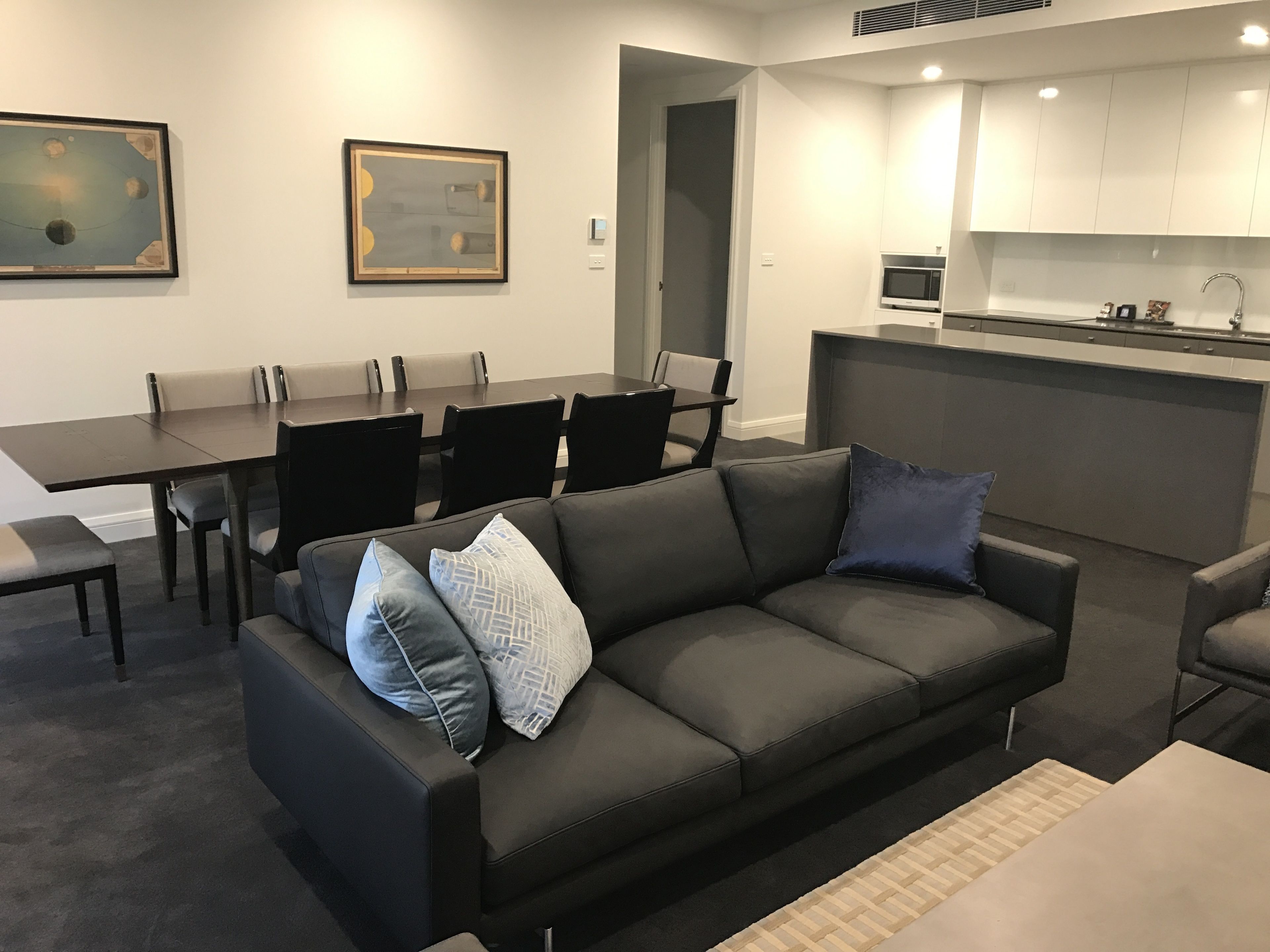 Una sala de estar moderna con un sofá gris oscuro, una mesa de comedor con sillas y una zona de cocina con armarios blancos y un microondas.