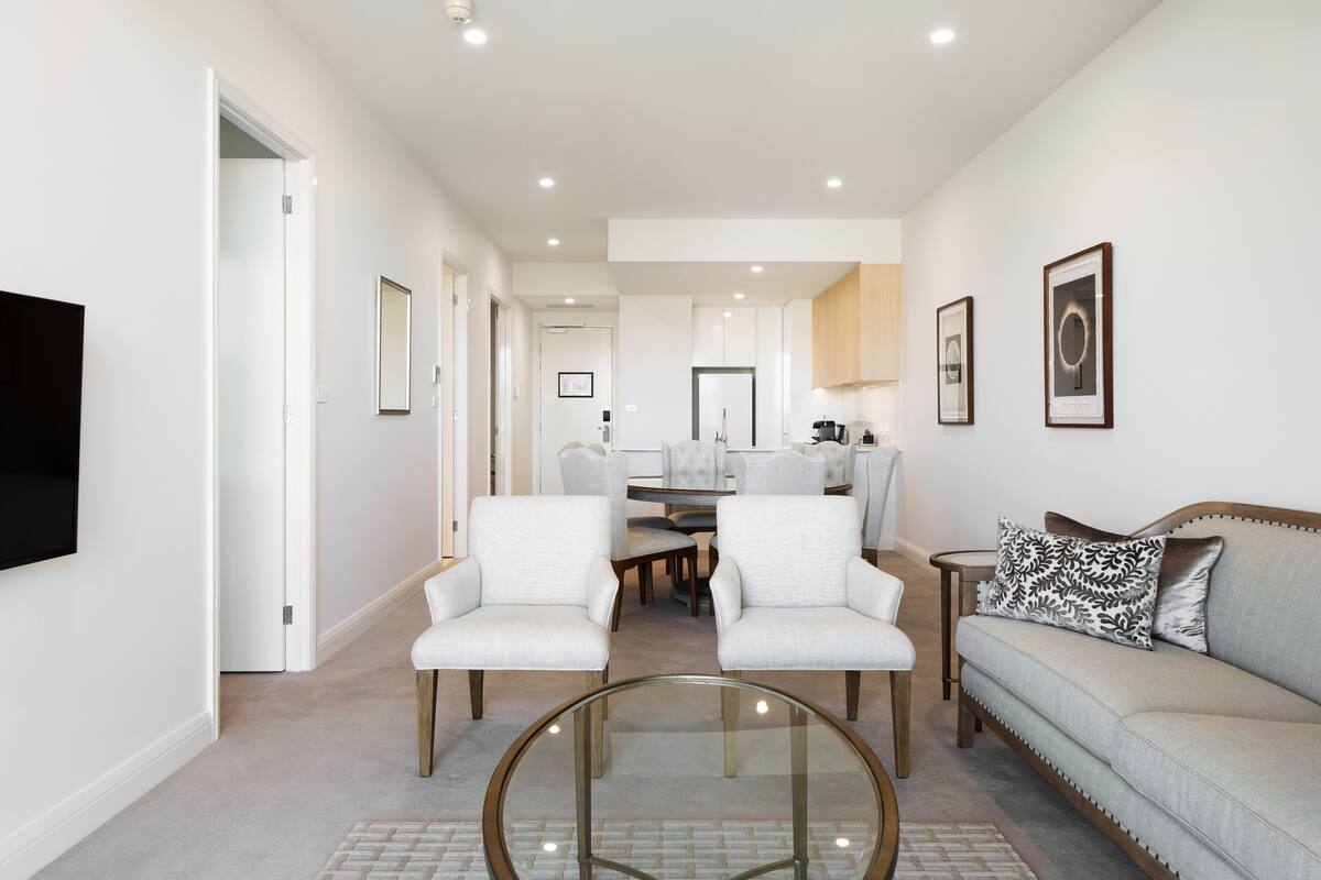 Una sala de estar moderna con una mesa de centro de vidrio, dos sillones blancos, un sofá gris y un comedor al fondo.