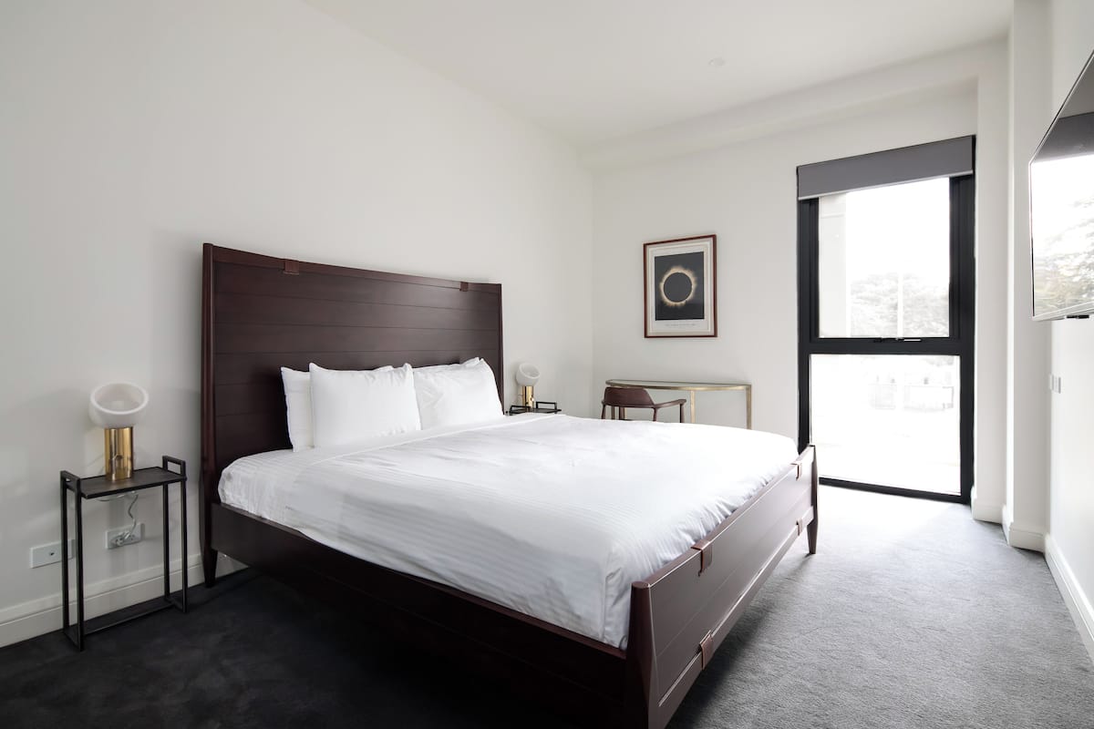 Una habitación de hotel moderna con una cama grande, un escritorio pequeño, una silla, un cuadro enmarcado en la pared y un ventanal amplio.