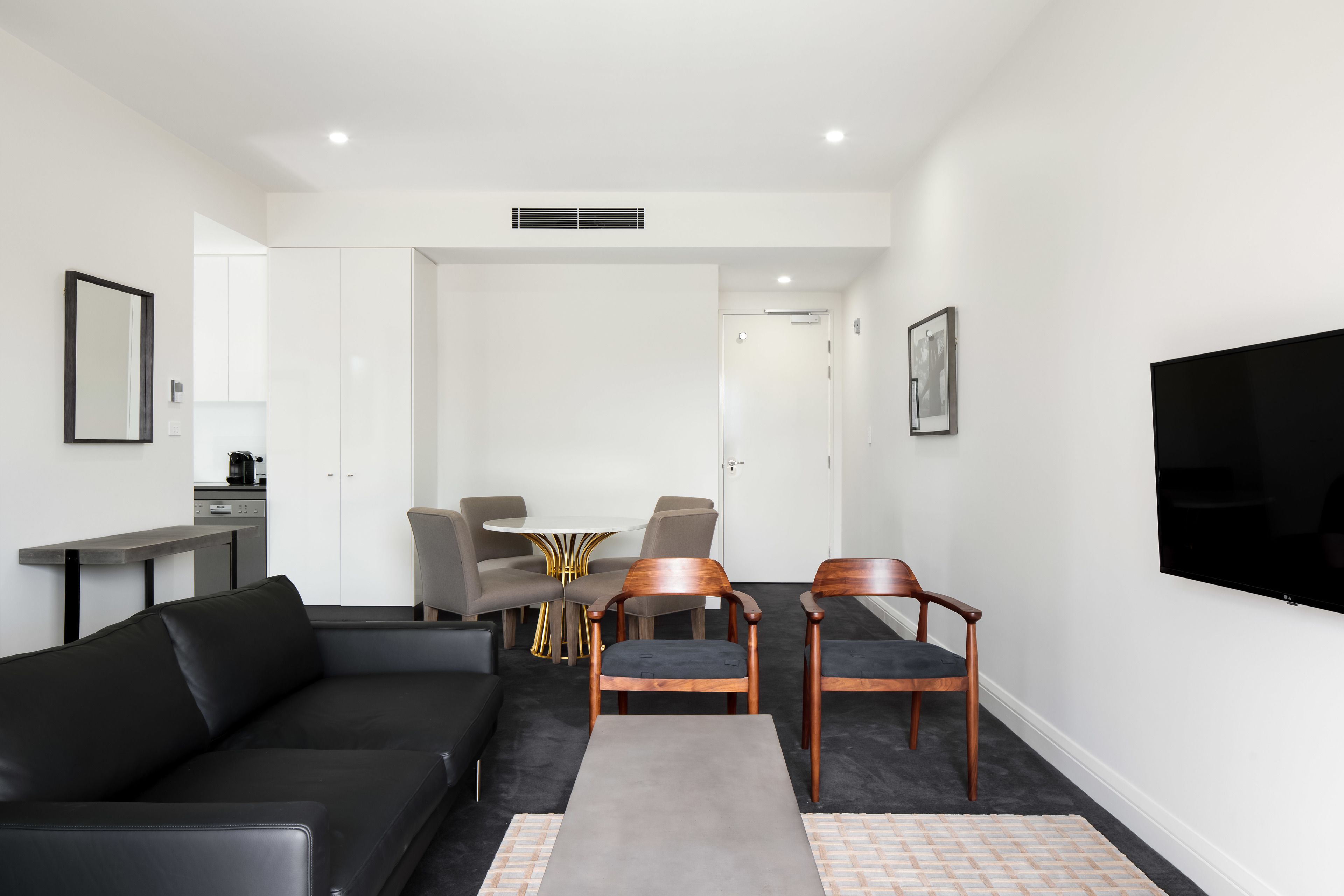 A modern living room with a black sofa, wooden chairs, a small table, and a flat-screen TV mounted on the wall.