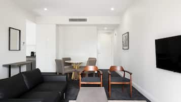A modern living room with a black sofa, wooden chairs, a small table, and a flat-screen TV mounted on the wall.