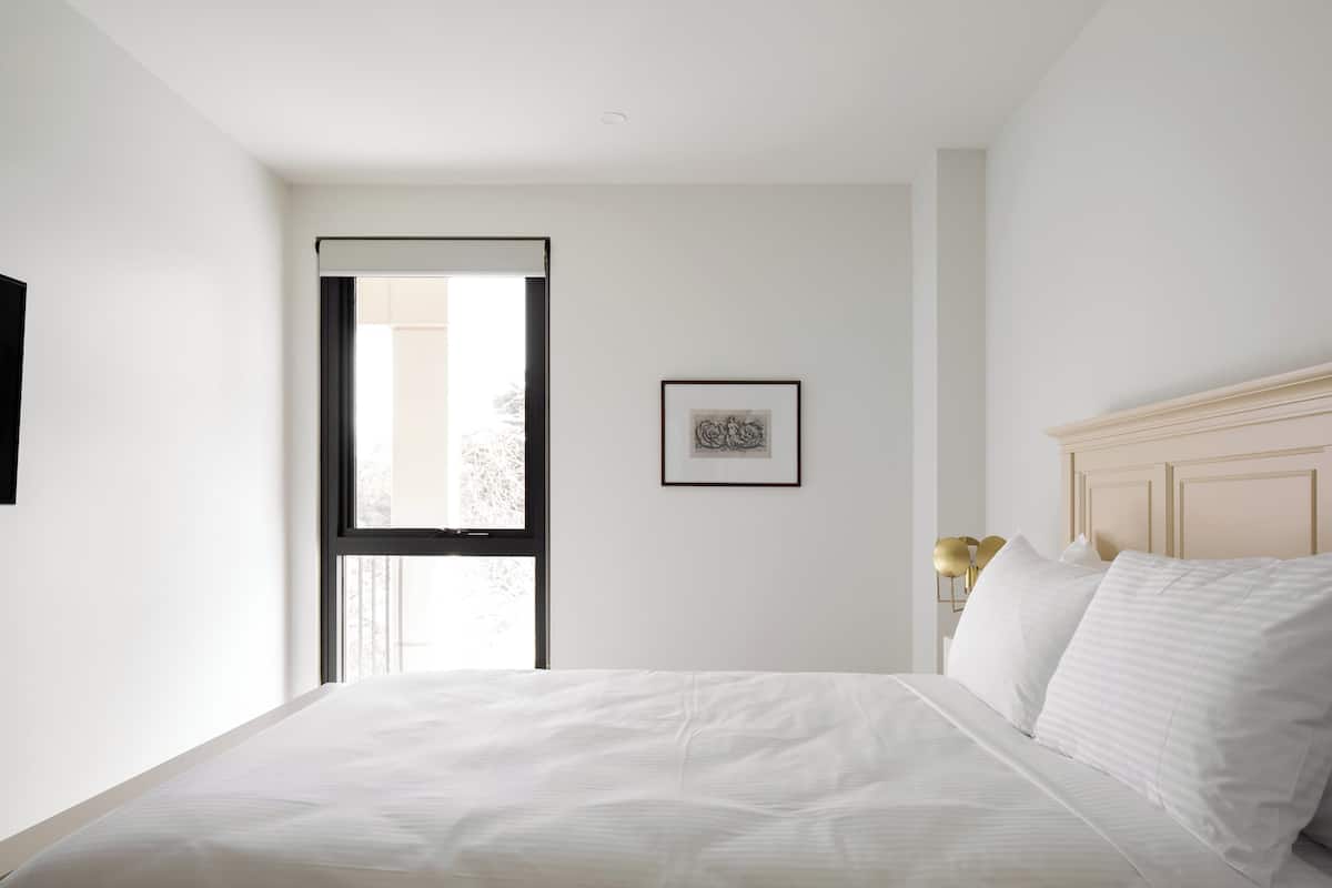 Un dormitorio con cama, ventana, un cuadro enmarcado y una lámpara de noche.