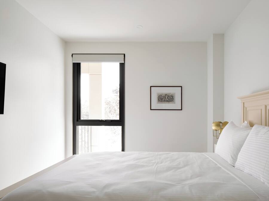 A bedroom with a bed, a window, a framed picture, and a bedside lamp.