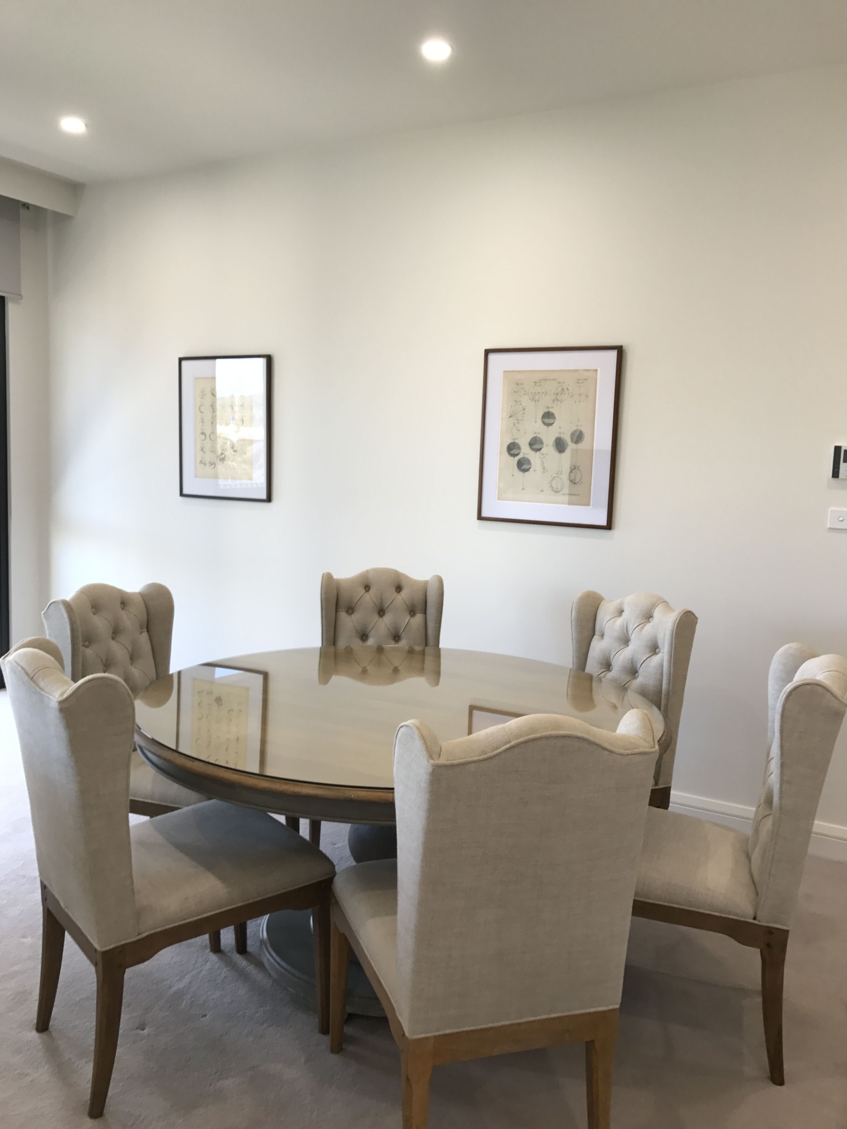 A round glass table surrounded by four tufted chairs with wooden legs. Two framed artworks are hung on the plain white walls.