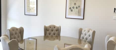 A round glass table surrounded by four tufted chairs with wooden legs. Two framed artworks are hung on the plain white walls.