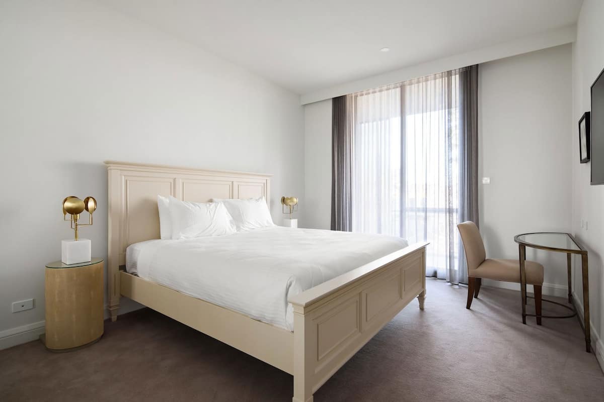 Un dormitorio ordenado con cama, mesitas de noche, una silla y televisor montado en la pared.