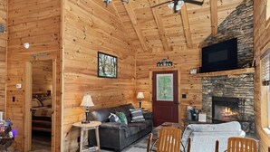 Interior de una cabaña con chimenea, paredes de madera, un sofá, una mesa de comedor y un televisor.