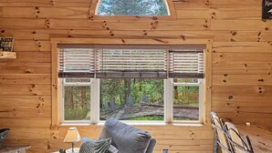 Interior de una cabaña de madera con una ventana grande, una chimenea, un sofá y una mesa de comedor.
