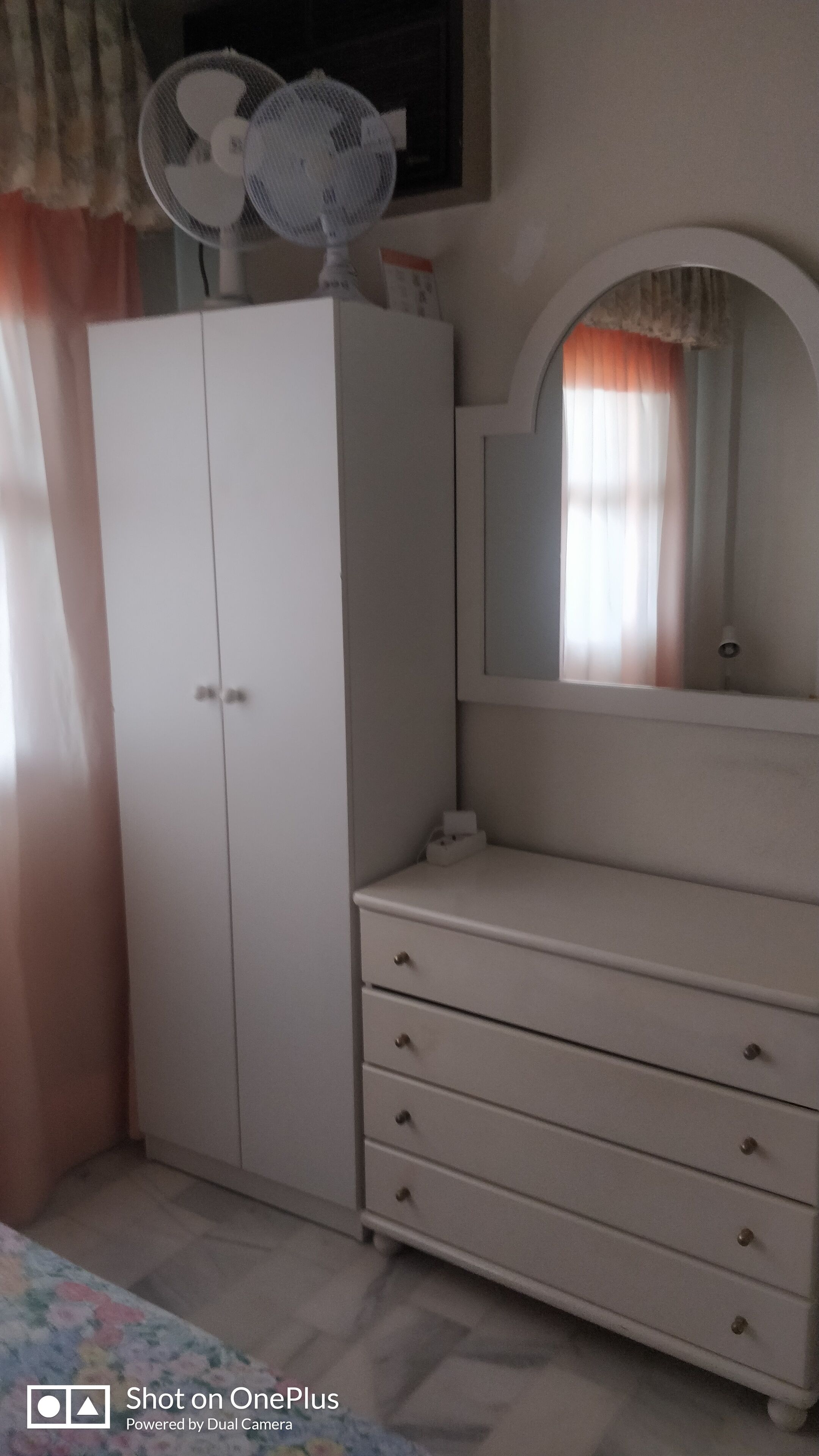 Une chambre avec une armoire blanche, une commode, un miroir et un ventilateur de plafond.