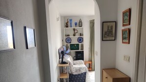 A hallway with a marble floor, a wooden cabinet, and a fan. There is a small table with a chair and a shelf with decorative items.