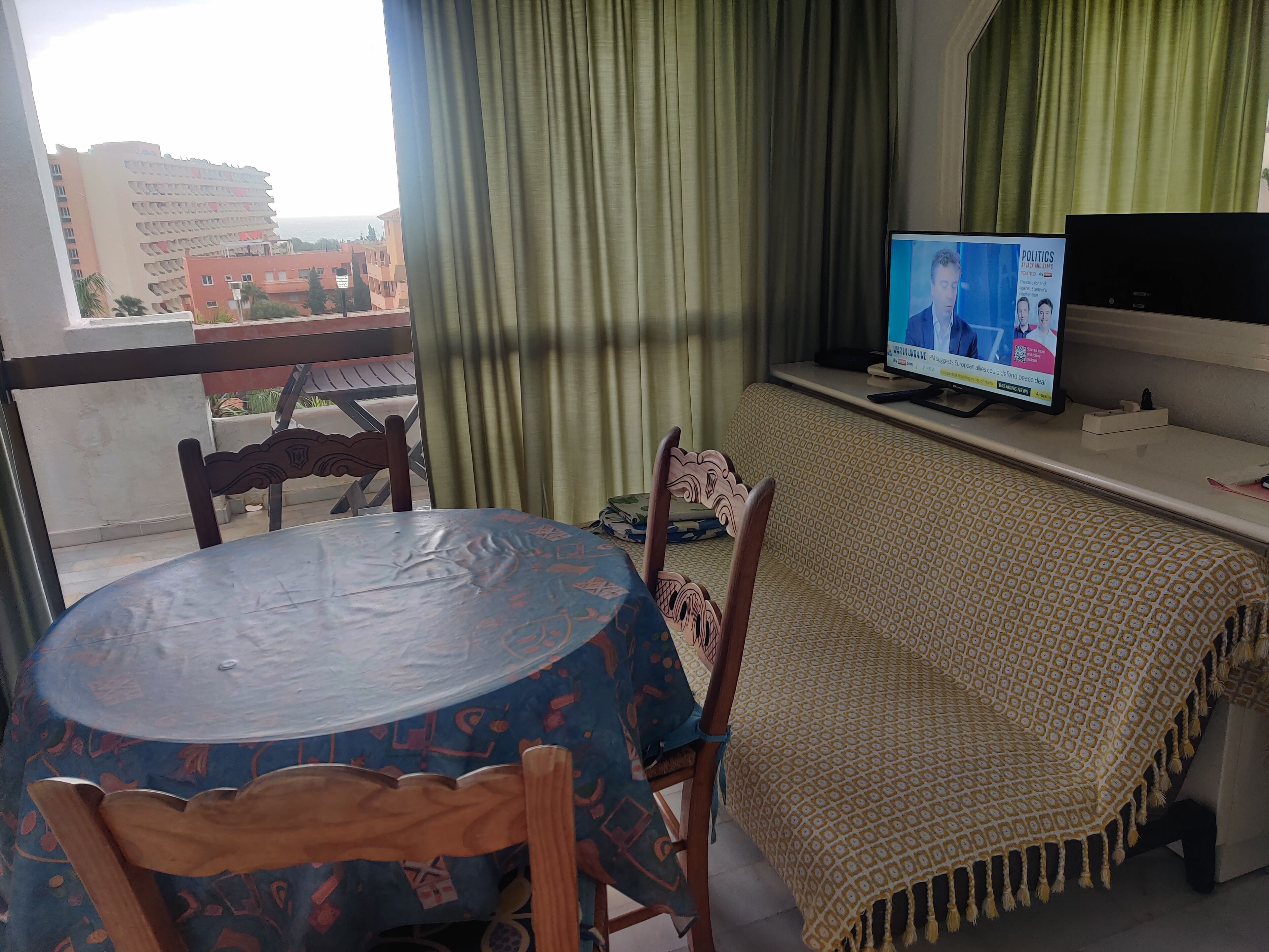 Une pièce avec une table ronde, deux chaises, un téléviseur posé sur un meuble, une nappe à motifs, un rideau vert et une vue sur les bâtiments à l’extérieur.
