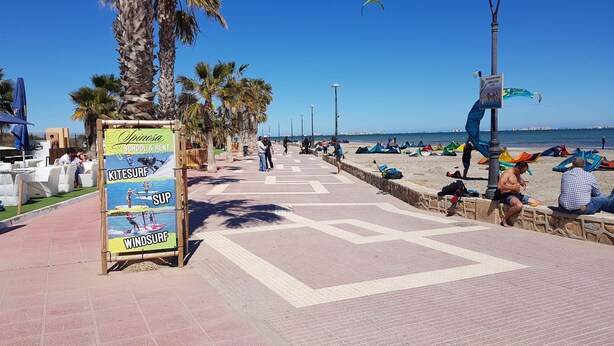 A beachside walkway with palm trees, a signboard for a school and surf rental, and beachgoers.