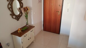 Un pasillo con una puerta de madera, un armario blanco con cajones, un espejo y un jarrón con flores.