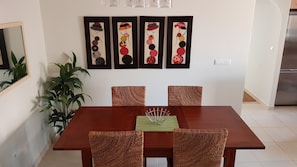 A dining area with a wooden table and four woven chairs. Above the table, there are four framed artworks with abstract designs. A potted plant is to the left of the table.