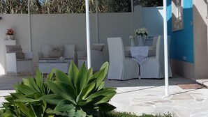 Un espace extérieur aménagé avec des meubles blancs, une table recouverte d’une nappe blanche et un pot de fleurs au premier plan.
