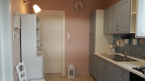 Une cuisine de taille réduite avec des meubles de couleur claire, une chaise blanche, une petite table blanche, une horloge au mur et une cuisinière.