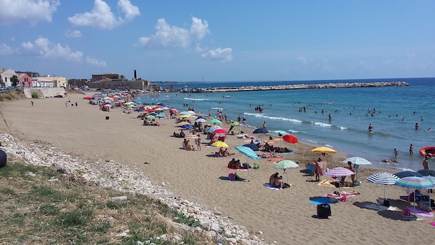 Une plage bondée où les gens se prélassent au soleil et se baignent, des parasols aux couleurs vives et une côte rocheuse.