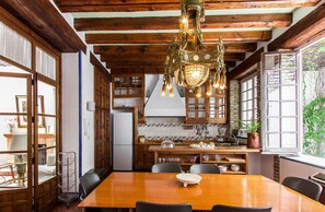 A spacious dining area with a wooden table and chairs, a chandelier, and a view of the kitchen and garden.