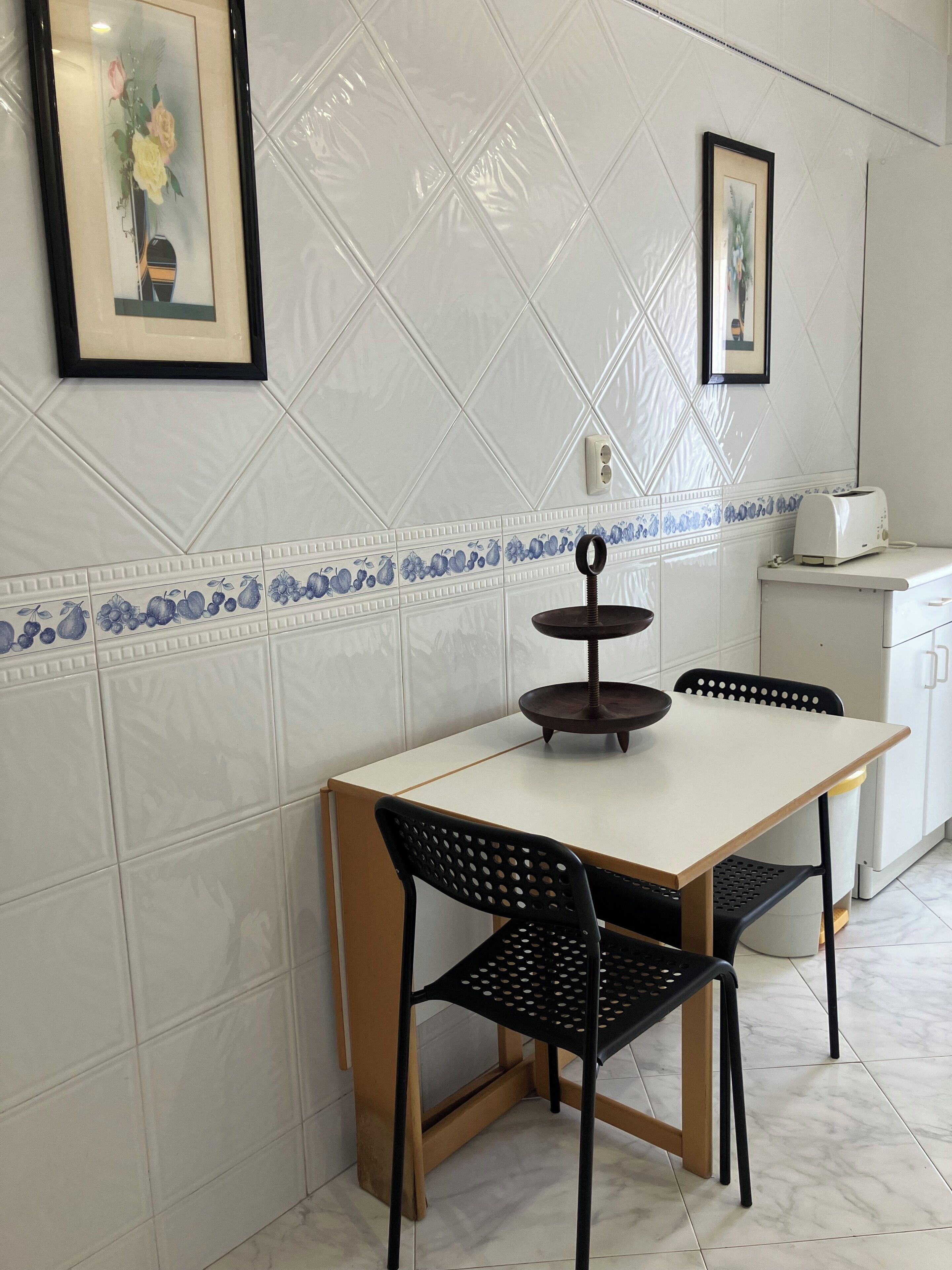 Un área de comedor con salpicadero de azulejos blancos, una mesa blanca, dos sillas negras y un expositor negro de tres niveles.