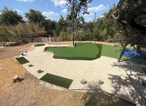 A landscaped outdoor area with a green artificial turf, a sandy path, and a wooden fence. - Bachelor  Party HQ, pickleball/golf, 16 beds. DT (Austin)