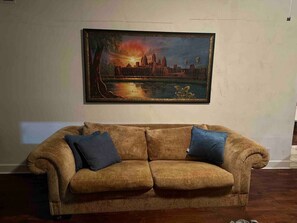 A room with a brown sofa, two blue cushions, and a framed painting of a sunset over a cityscape. - Bachelor Pad 18 beds, Golf, Pool, Pickleball, 5min to 6th street (Austin)