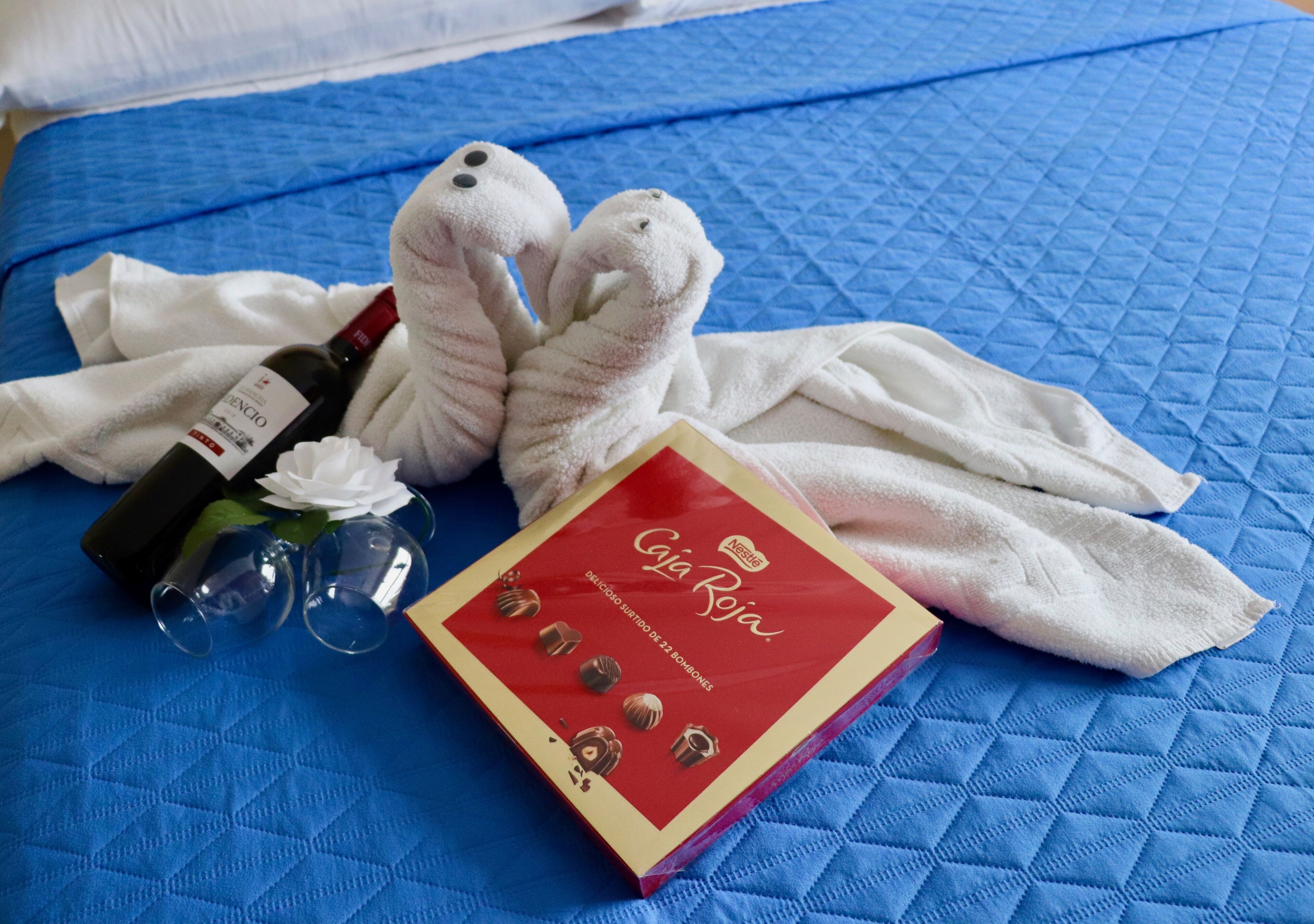 Un lit recouvert d’une couverture matelassée bleue, une bouteille de vin, deux peluches, une serviette blanche et une boîte de biscuits Tollhouse de la marque Nestlé.
