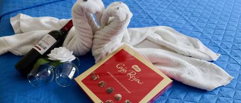 Un lit recouvert d’une couverture matelassée bleue, une bouteille de vin, deux peluches, une serviette blanche et une boîte de biscuits Tollhouse de la marque Nestlé.