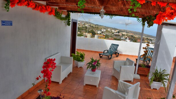 A covered outdoor seating area with white wicker furniture, potted plants, and a view of the town in the background. - CASA YOEL "PRIVACY, WARMTH, GUARANTEED REST, IDEAL FOR FAMILIES, NEW !!! (Granadilla)