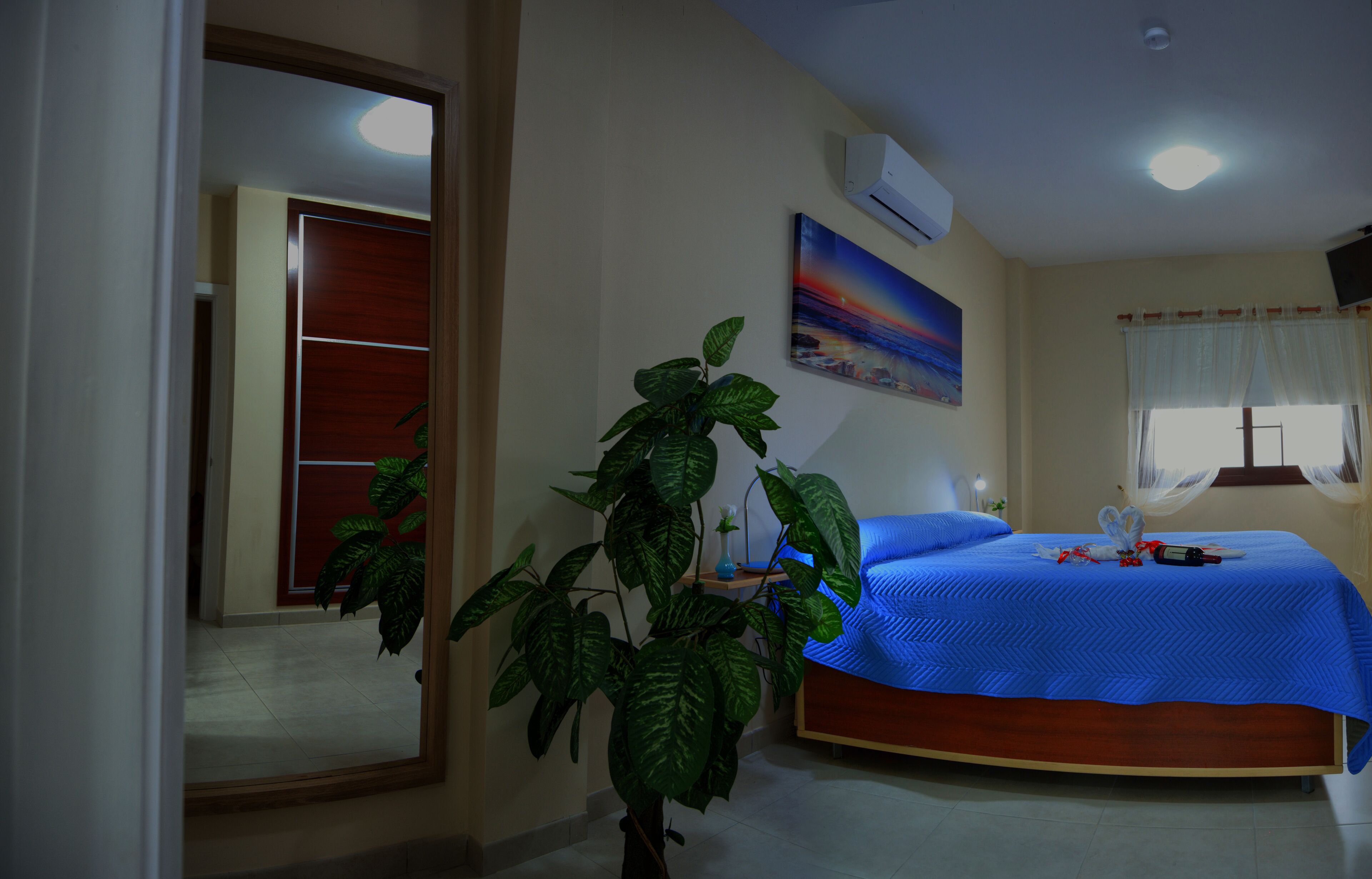 A bedroom with a bed, a potted plant, a painting, and a window with curtains.
