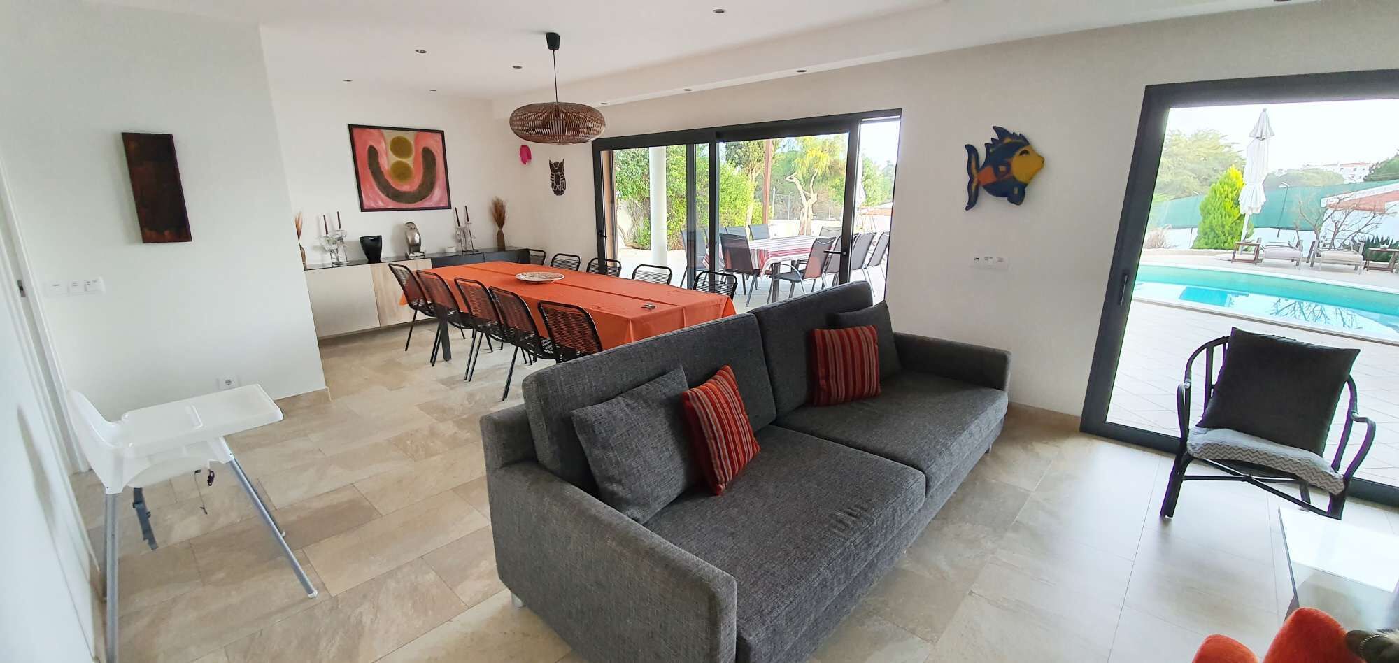 A modern living room with a grey sofa, a dining area with a red table, and a view of a swimming pool.