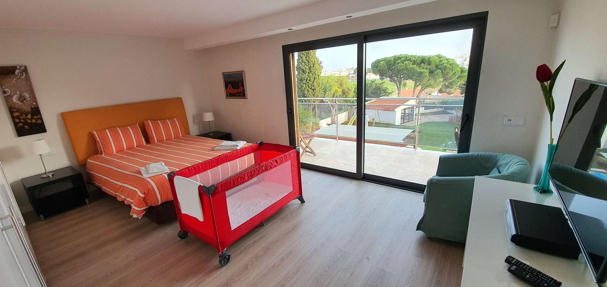 Une chambre d’hôtel moderne avec un lit, un parc pour enfants rouge, une chaise verte, une télévision et un balcon offrant une vue sur les arbres et les bâtiments.