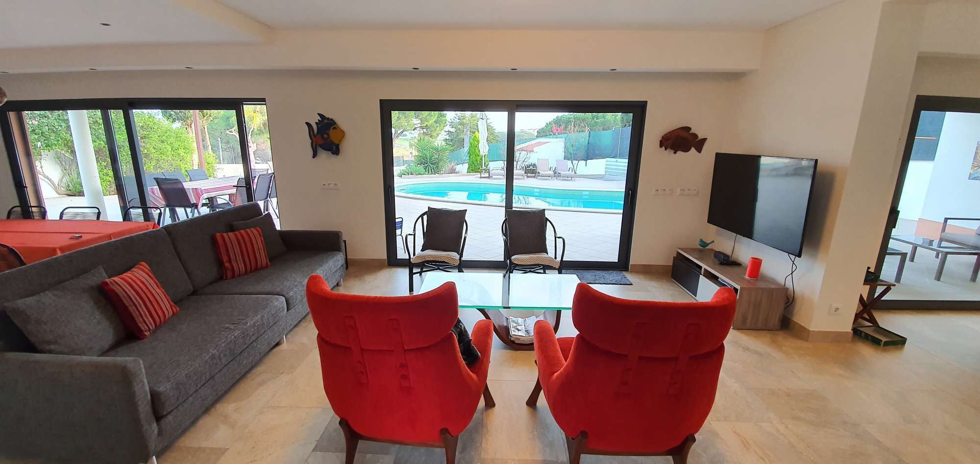Un salon spacieux avec un canapé gris, des fauteuils rouges, un téléviseur et une vue sur une piscine, visible à travers de grandes portes coulissantes.