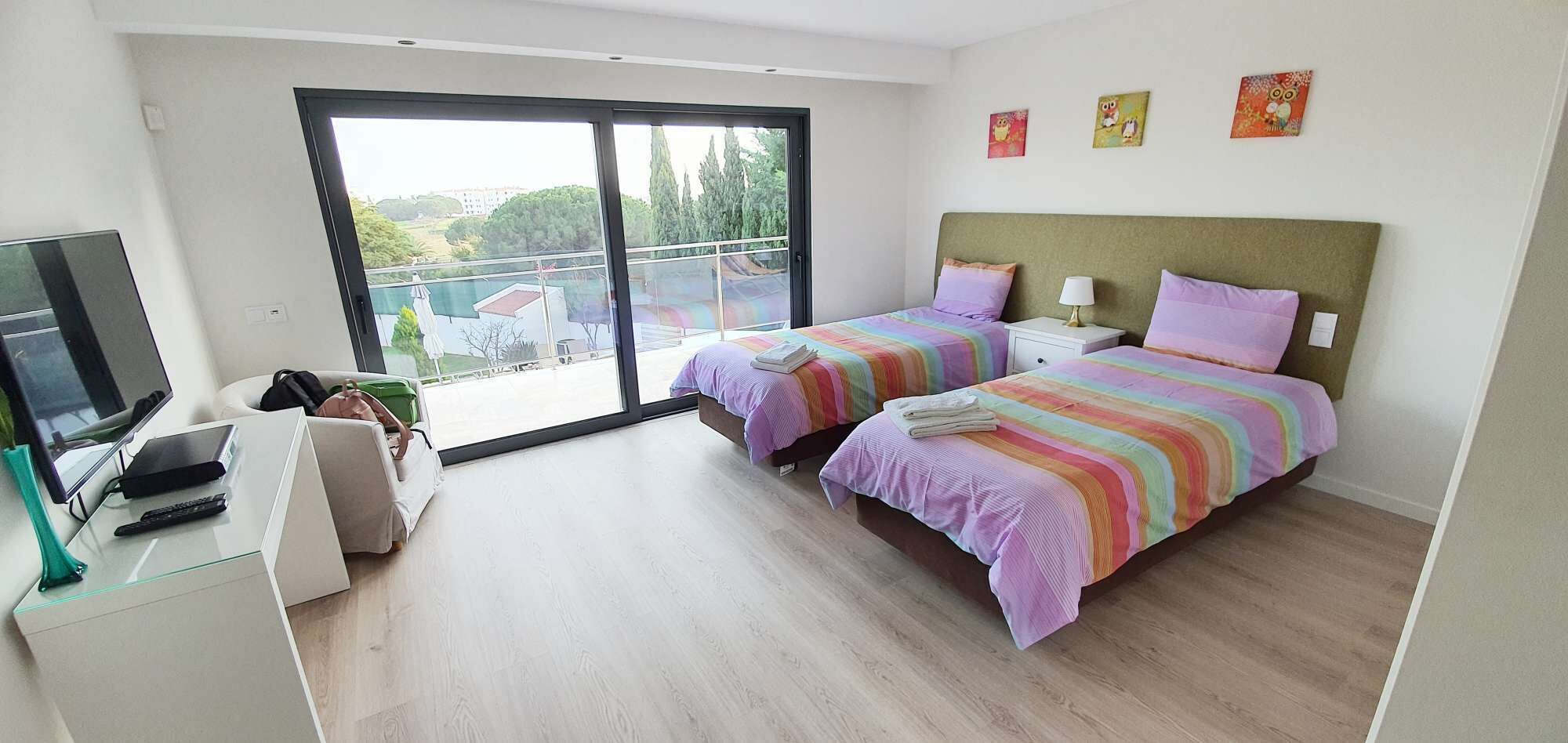 A modern bedroom with two beds, a TV, and a balcony with a view of buildings and trees.
