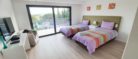 A modern bedroom with two beds, a TV, and a balcony with a view of buildings and trees.