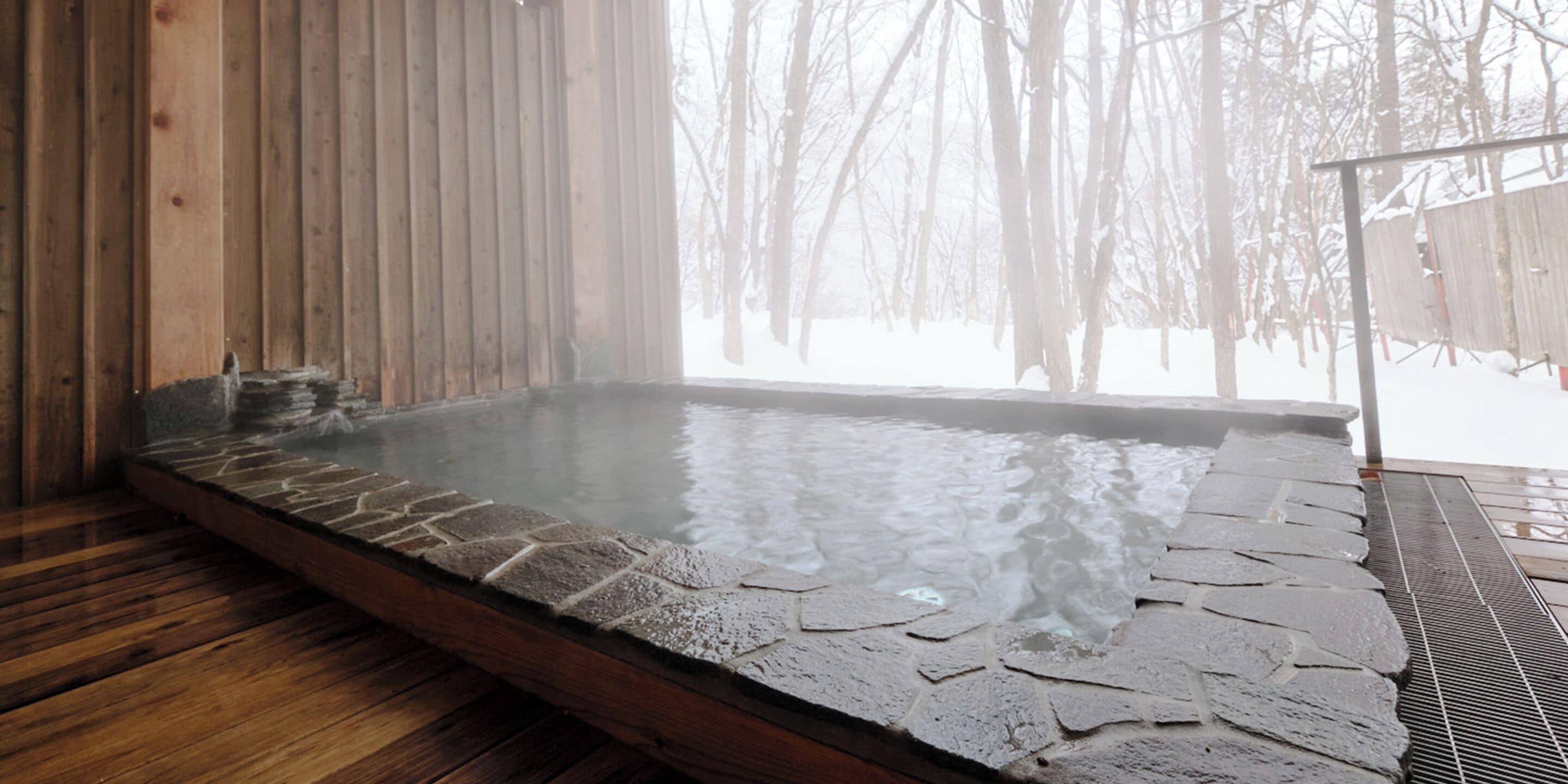 A hot spring bath with stone flooring, wooden walls, and a view of a snowy forest.